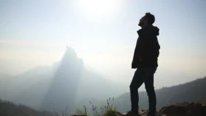 A person standing at the base of a mountain, looking upward with determination and calm confidence, misty peaks in the distance, morning light breaking through clouds, serene landscape background