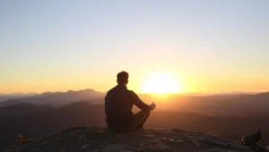 Person sitting on a mountain overlook at sunrise in Colorado, looking peaceful and focused, mountains in background, natural lighting, contemplative pose