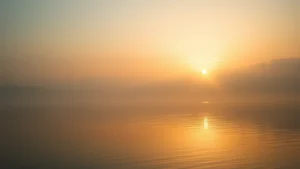 Serene sunrise over calm water, soft golden light reflecting on surface, peaceful natural landscape with mist rising, minimalist composition suggesting hope and new beginnings